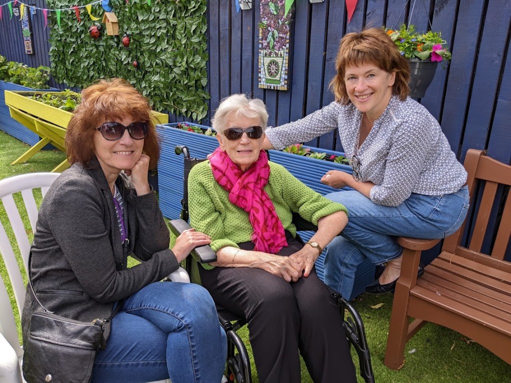 A lady in a wheelchair, flanked by two female visitors.