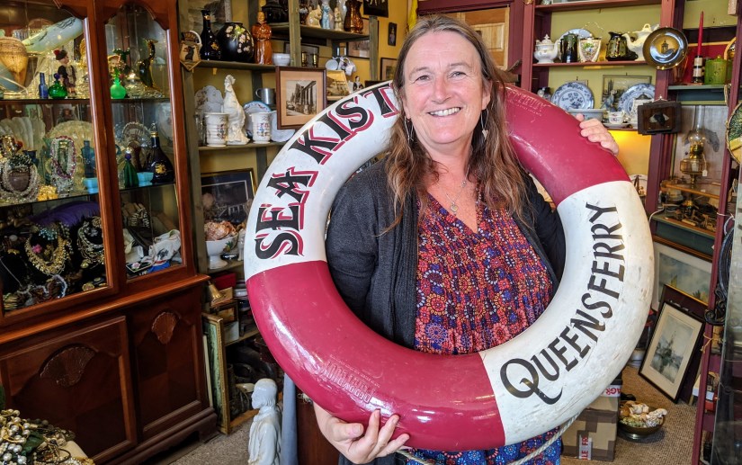 Woman holding a life buoy with the words Sea Kist Queensferry on it, in an antiques shop.